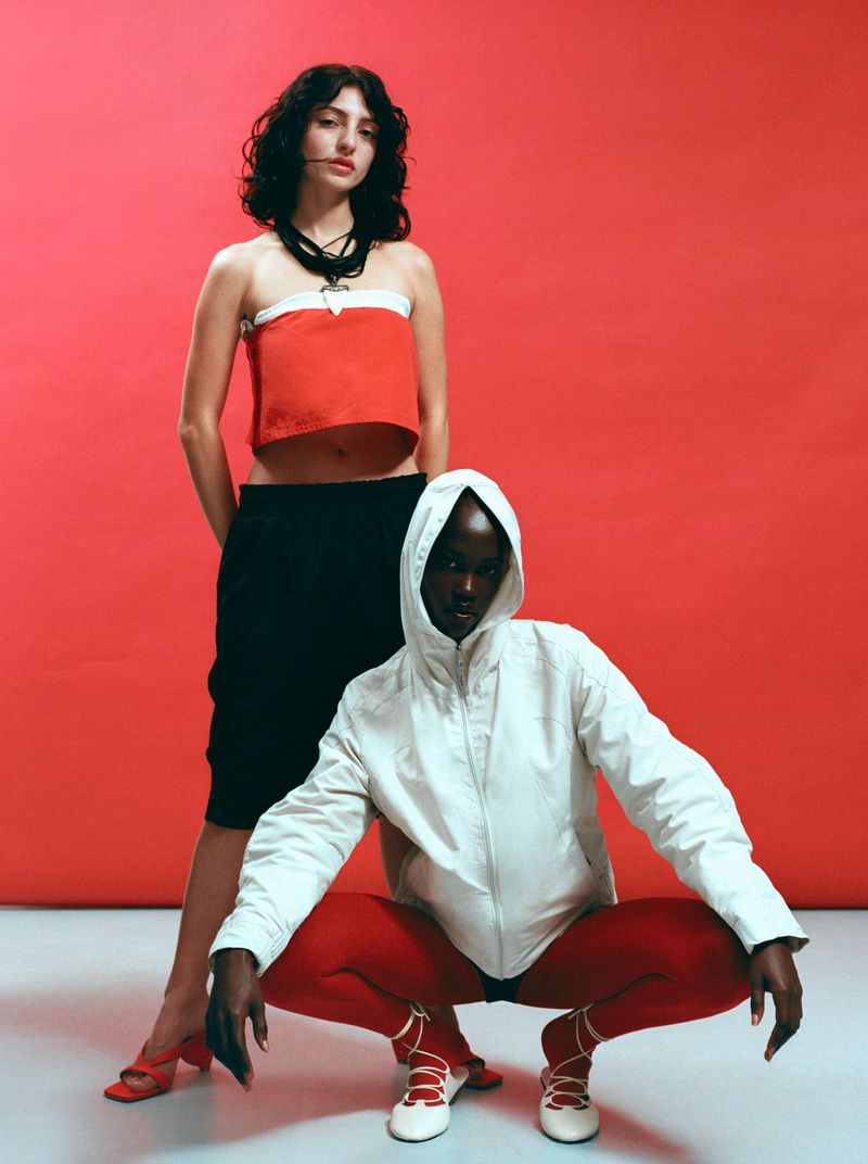 Two fashion models posing against a bright red backdrop, one standing and one crouching in a white hooded jacket.