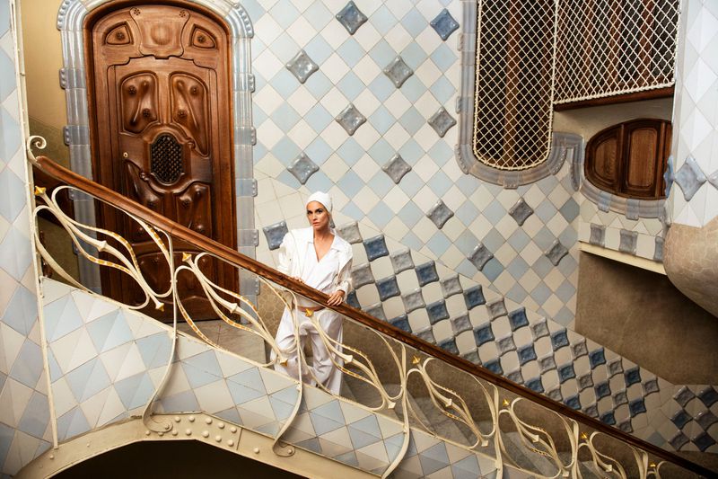 A woman in a white hooded jumpsuit stands on an ornate staircase with ceramic tiled walls and a carved wooden door.