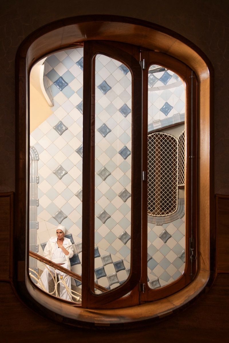 Model in white attire seated near tiled walls framed through an arched wooden window for Harper's Bazaar España editorial