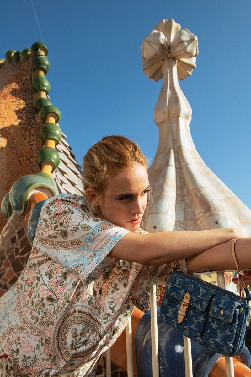 A woman in a patterned dress leans over a railing on a Gaudi rooftop next to a blue denim monogram shoulder bag under a clear sky.