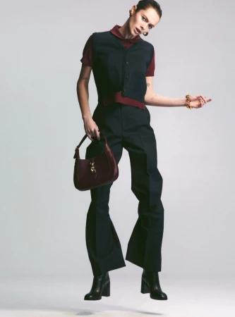Fashionable woman in a black vest and pants, posing mid-air while holding a burgundy purse, with black heeled boots on a grey backdrop.
