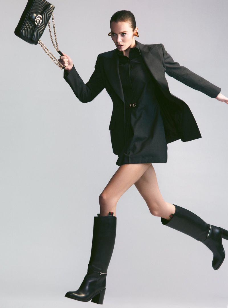 Emma Boyd leaps across a grey studio backdrop wearing a tailored black blazer, knee-high boots, and holding a quilted chain handbag.