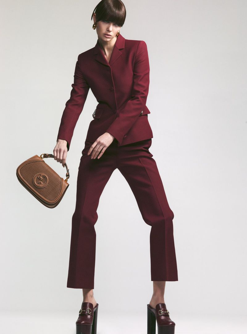 A stylish adult woman in a maroon suit with retro makeup, holding a Gucci handbag, and wearing platform heels against a white backdrop.