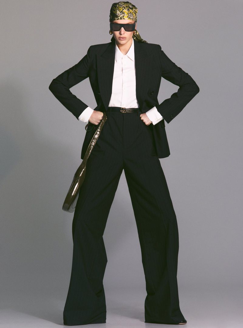 A woman in a black suit and patterned headscarf poses in a studio, creating a high-fashion editorial image with neutral gray backdrop.
