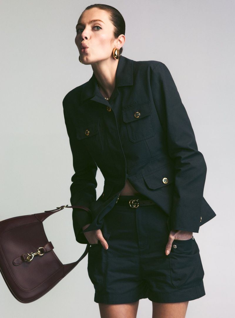 Brunette woman in dark jacket, shorts, and gold earrings, pouting with a leather purse on a neutral background in a studio shot.