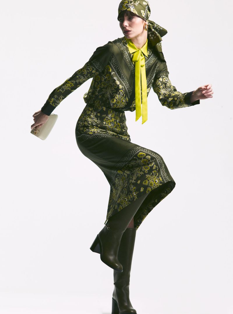 A woman in a patterned green outfit and matching headscarf, accented with a yellow shirt and boots, posing on a white backdrop.