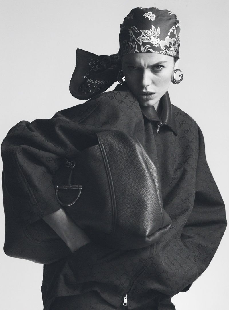 Model Emma Boyd posing in a printed headscarf, oversized zip jacket, and large hoop earrings holding a black leather bag.