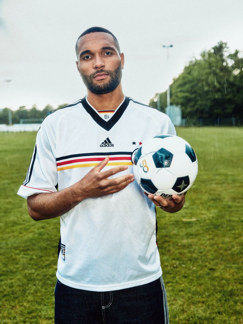 Jonathan Tah wearing a white soccer jersey holding a football on a green grass field for GQ Germany.