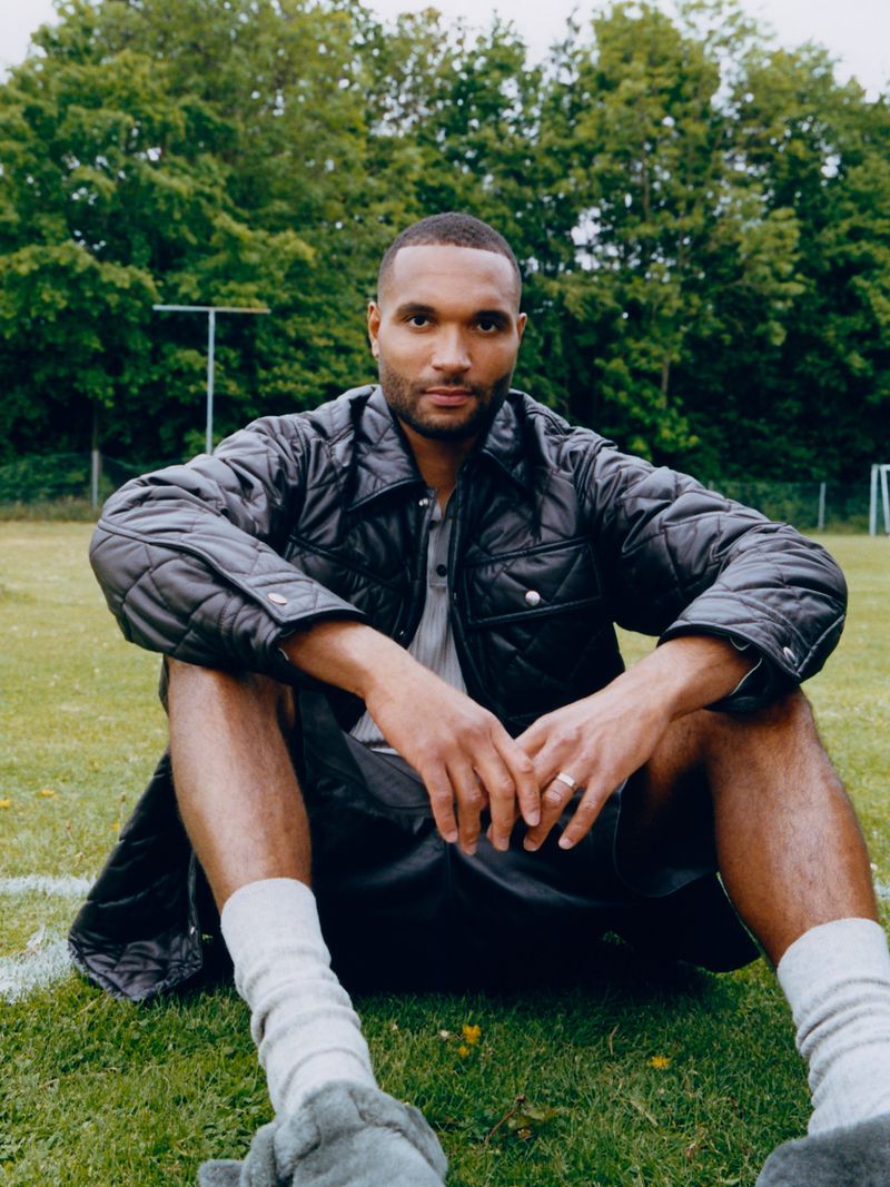Footballer Jonathan Tah sitting on a grassy field wearing a black quilted jacket and grey socks.