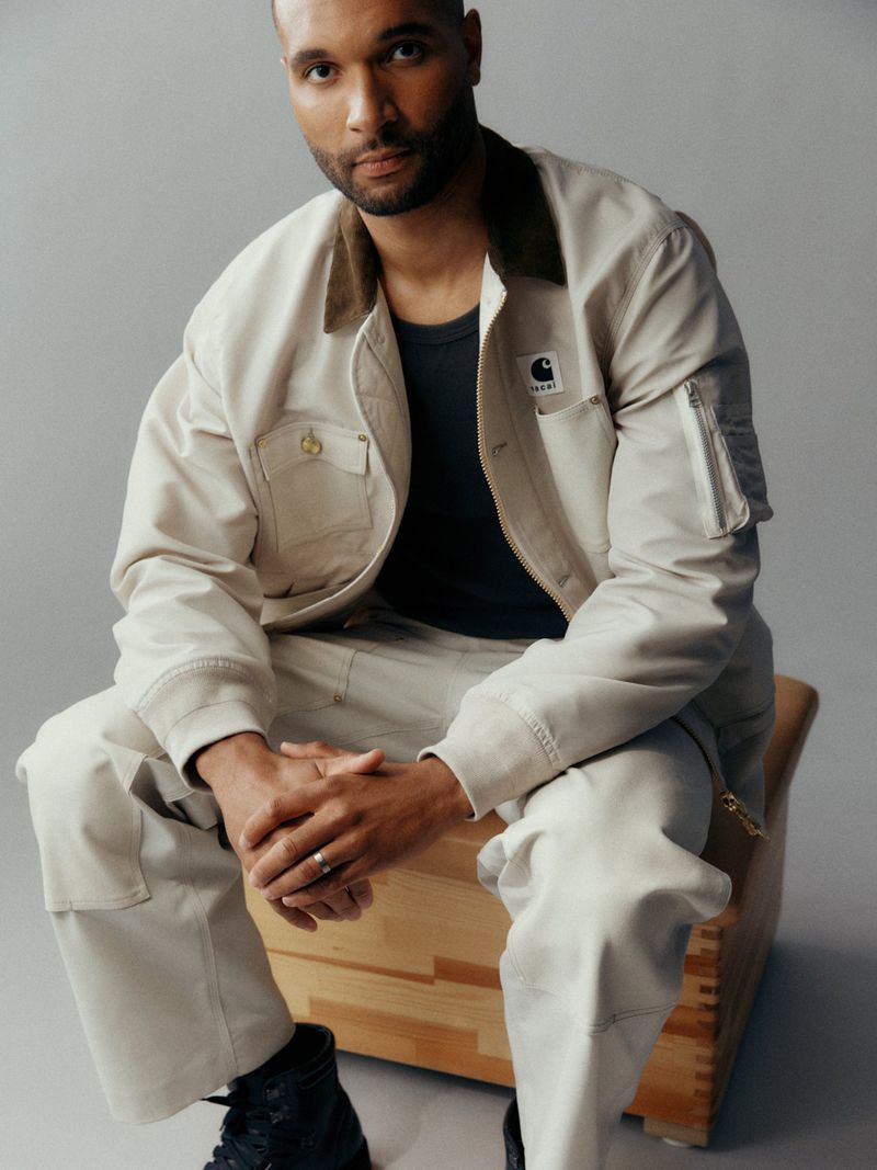 Jonathan Tah seated on a wooden box wearing an off-white workwear jacket and matching pants against a neutral grey studio backdrop.
