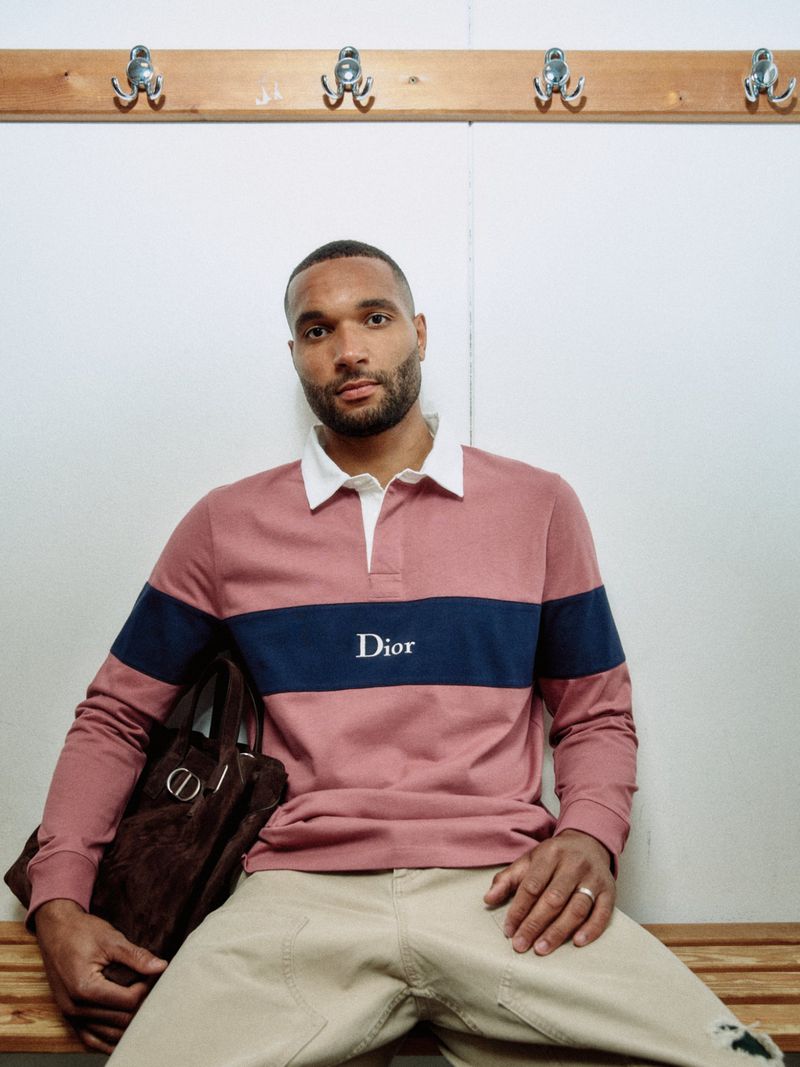 Jonathan Tah seated on a bench wearing a dusty rose Dior polo shirt with a navy stripe and beige trousers.