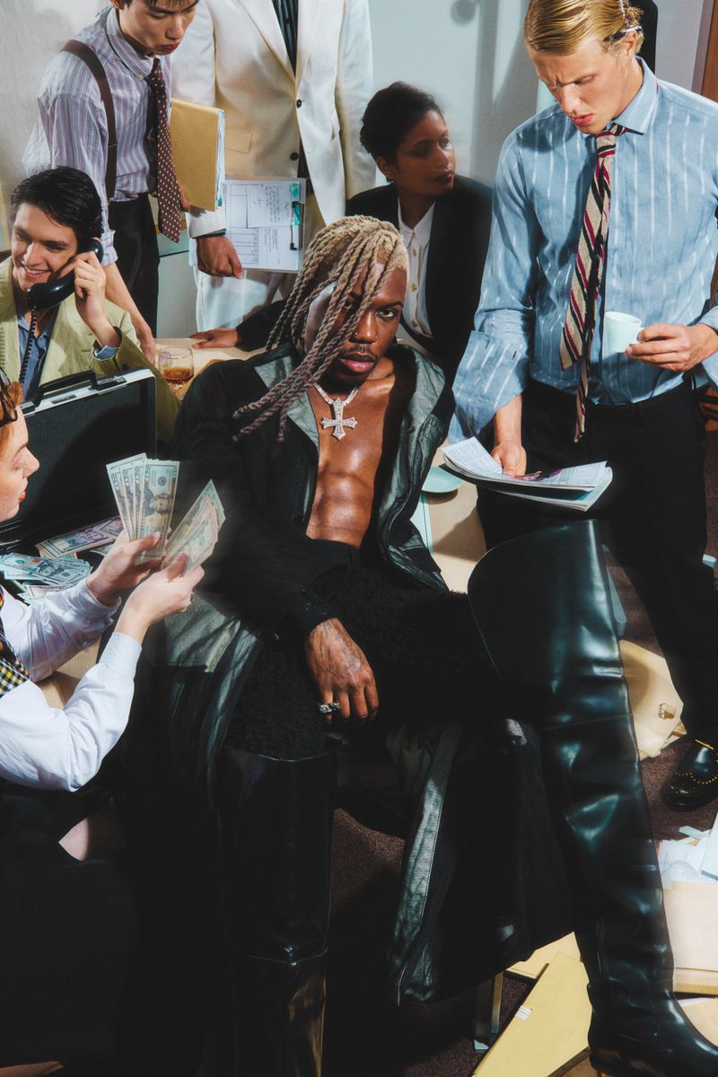 A man sits in a black coat and tall boots, surrounded by office workers holding papers and money in an indoor office setting.