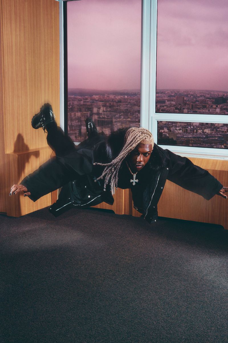 An adult male wearing black clothing poses in a room with a city view. He is captured mid-motion in an unconventional pose.