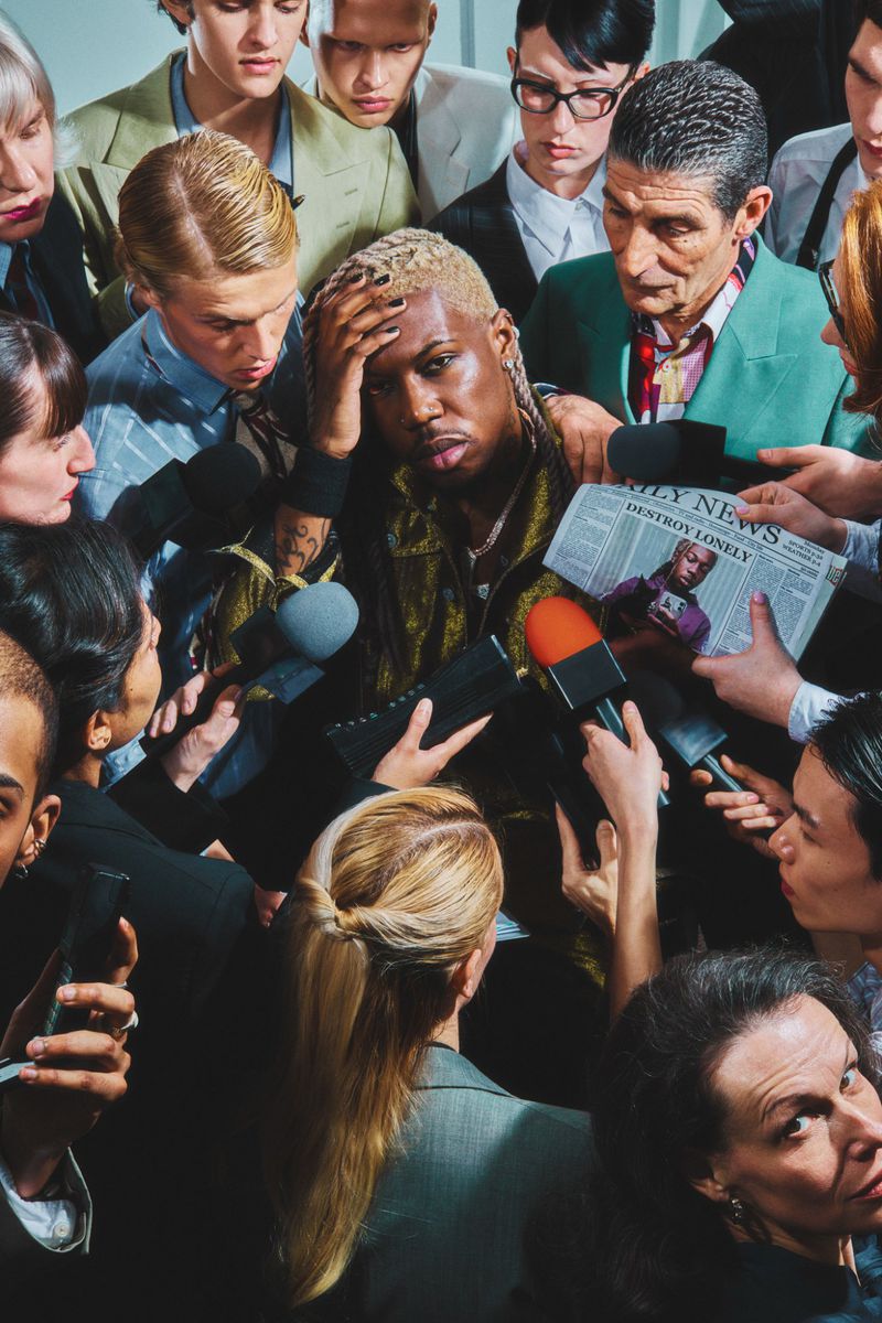 A group of reporters surround and interview a stylish adult male with platinum blonde hair, wearing a patterned jacket and jewelry.