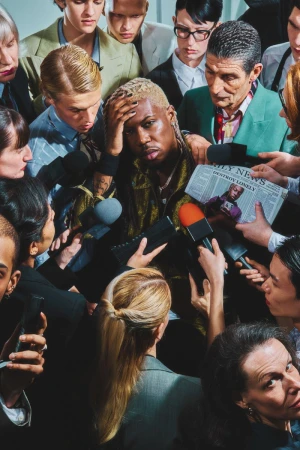 A group of reporters surround and interview a stylish adult male with platinum blonde hair, wearing a patterned jacket and jewelry.