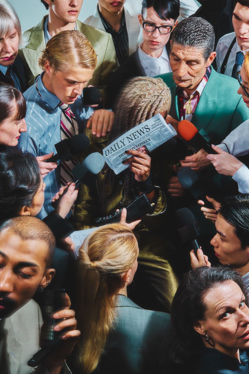 Person reads 'Daily News' newspaper surrounded by reporters holding microphones in an indoor setting with a group of people.