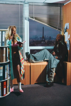 Woman with notepad and man in window with Eiffel Tower backdrop. Woman poses next to shelves of binders, the man in the window.