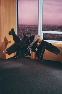 Man with braided blonde hair in a black jacket suspended mid-air inside a room with large windows overlooking a city.
