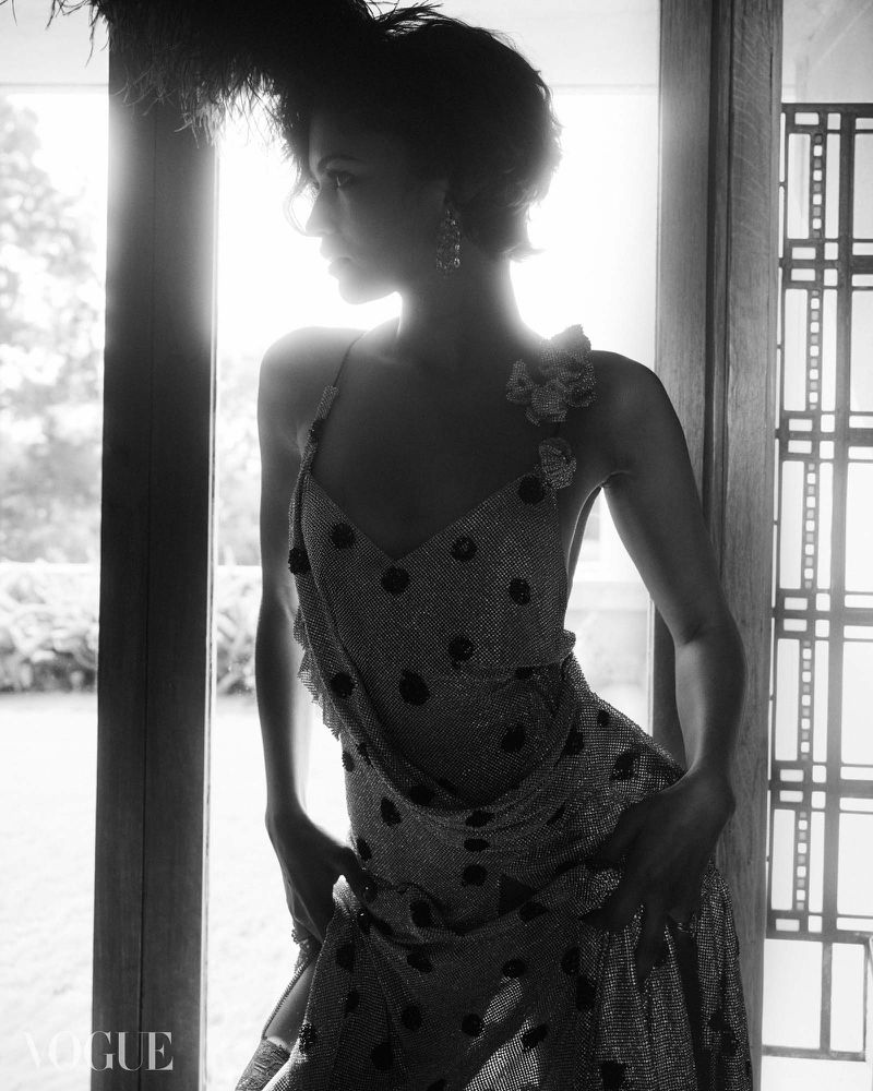 Zendaya posing in a polka dot slip dress and feather headpiece backlit in a doorway in black and white.