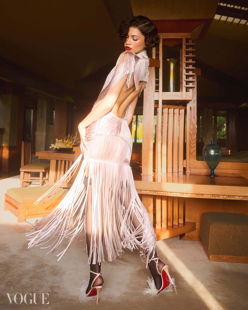 Zendaya posing in a backless blush fringe dress and stiletto heels inside a sunlit wooden architectural interior.