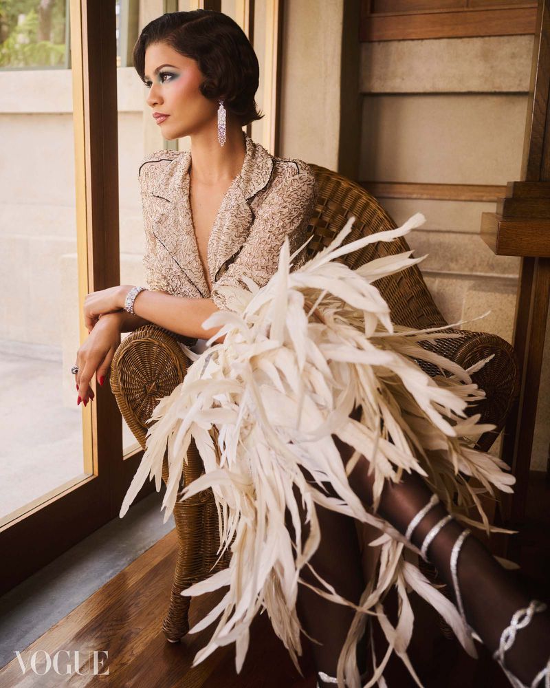 Zendaya seated in a wicker chair wearing a tweed jacket, dramatic white feather skirt, and strappy heels next to a window.