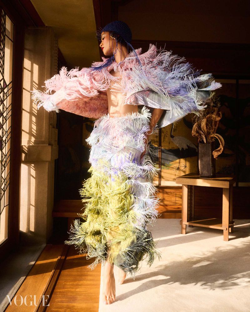 Zendaya poses indoors wearing a multi-tiered pastel feathered fringe cape dress and knit cap in warm sunlight.