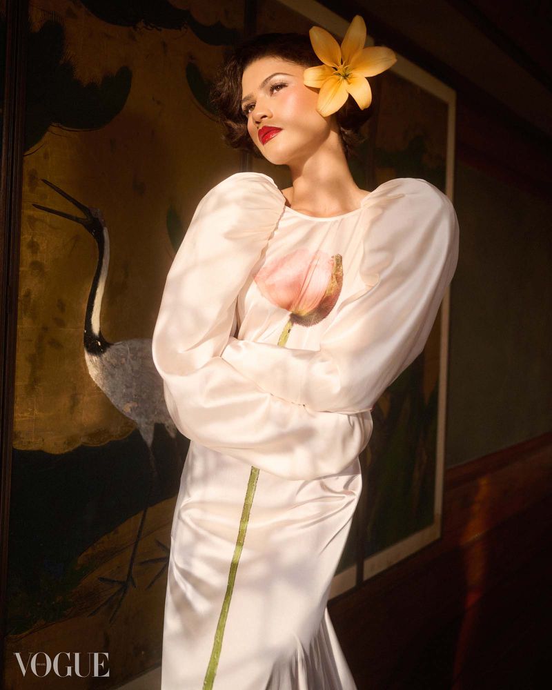 Zendaya wears a white puff-sleeve satin dress with a printed pink flower and a yellow lily in her hair before a painted screen.