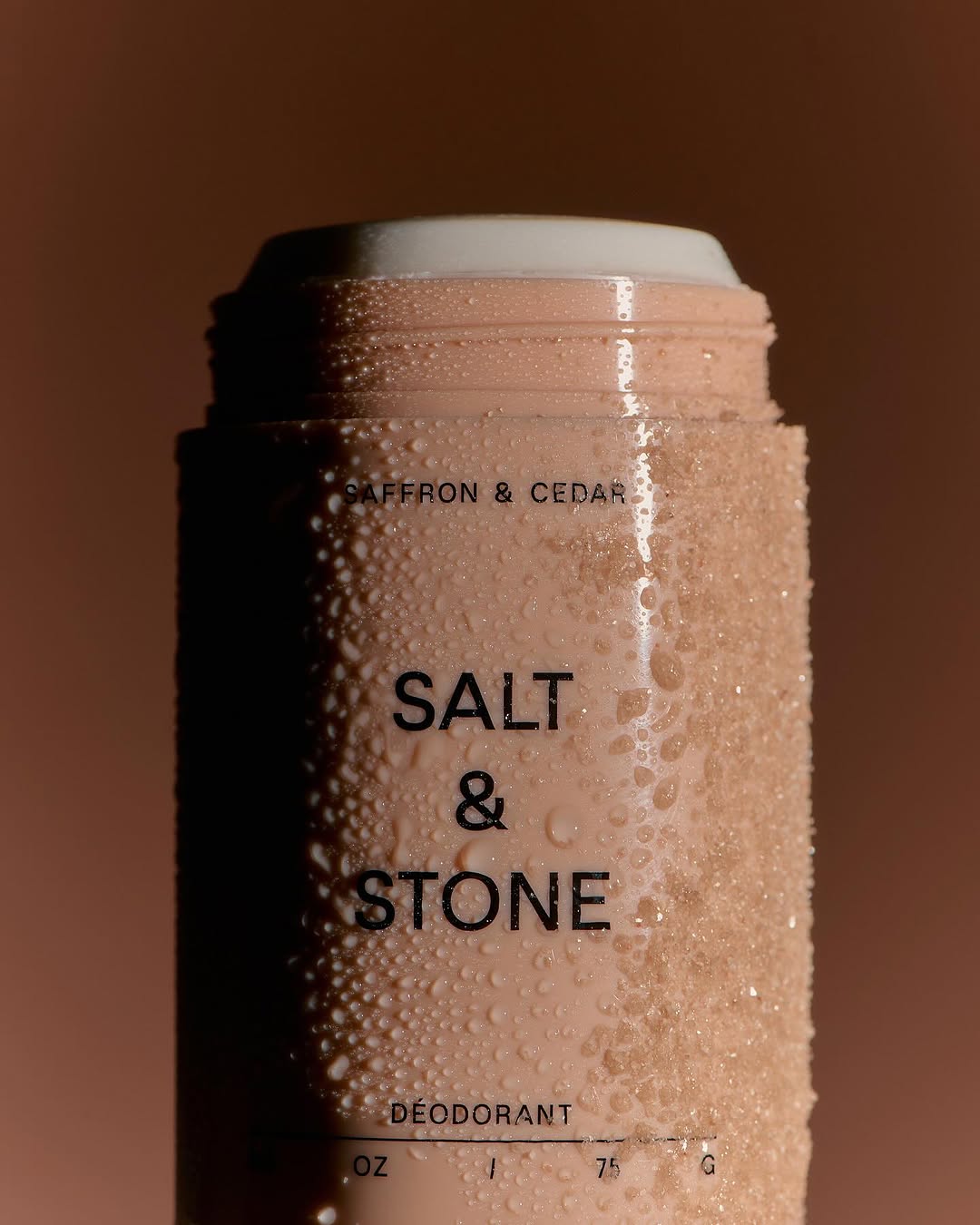 Close-up of a Salt & Stone deodorant container against a blurred background, featuring water droplets on the container's surface.