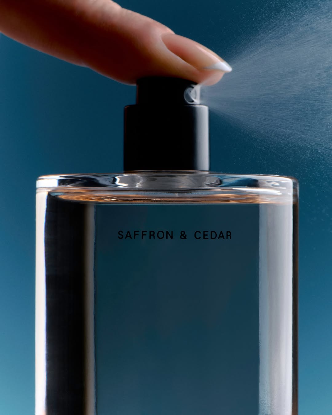 Close-up of a finger pressing the spray nozzle of a clear glass perfume bottle against a blue background, releasing a mist.