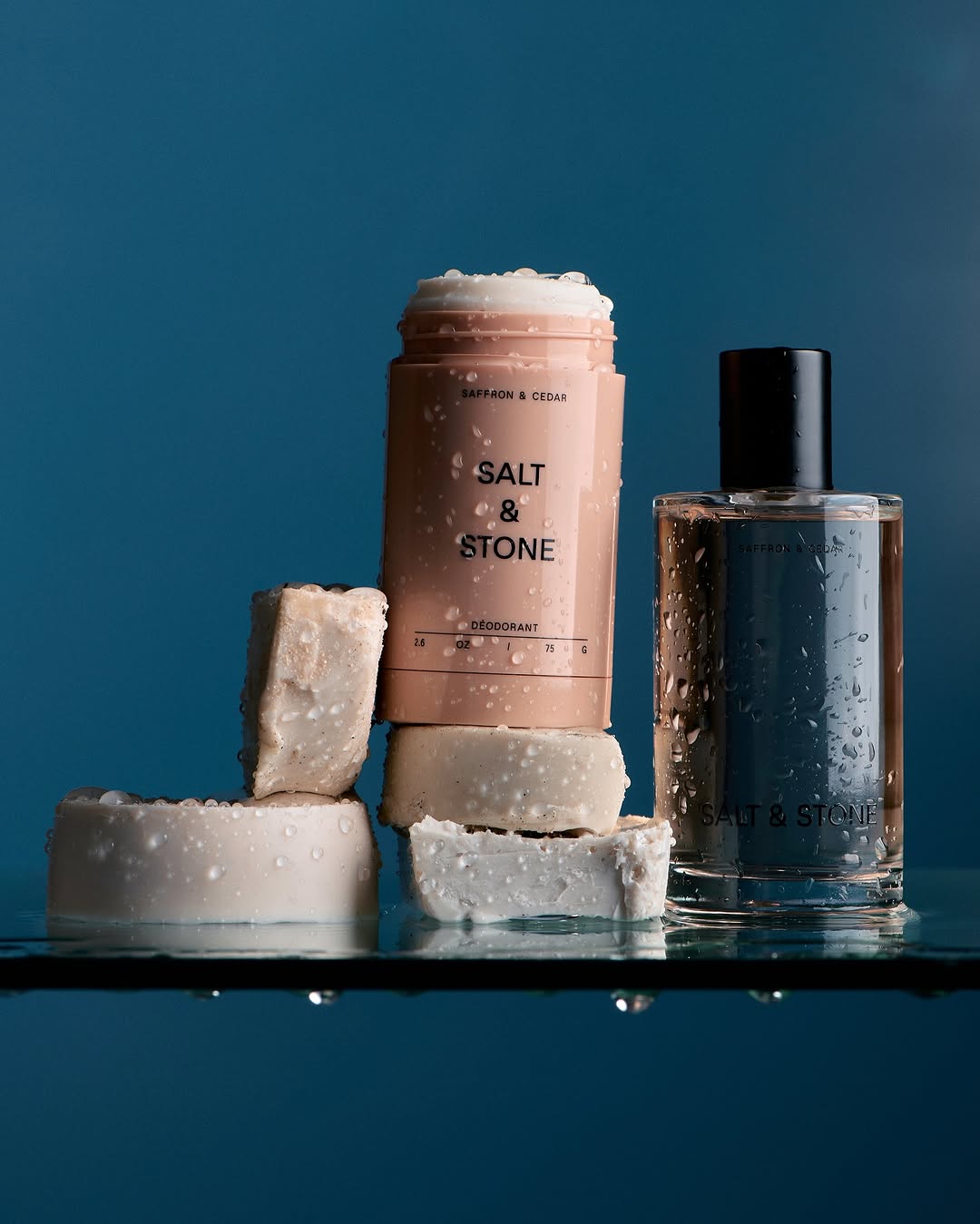 Close-up studio shot of Salt & Stone brand deodorant, perfume bottle, and cubed soaps on a wet glass surface against a blue background.