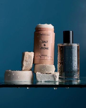Close-up studio shot of Salt & Stone brand deodorant, perfume bottle, and cubed soaps on a wet glass surface against a blue background.