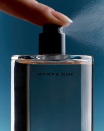 Close-up of a finger pressing the nozzle of a Salt & Cedar perfume bottle, releasing a fine mist spray