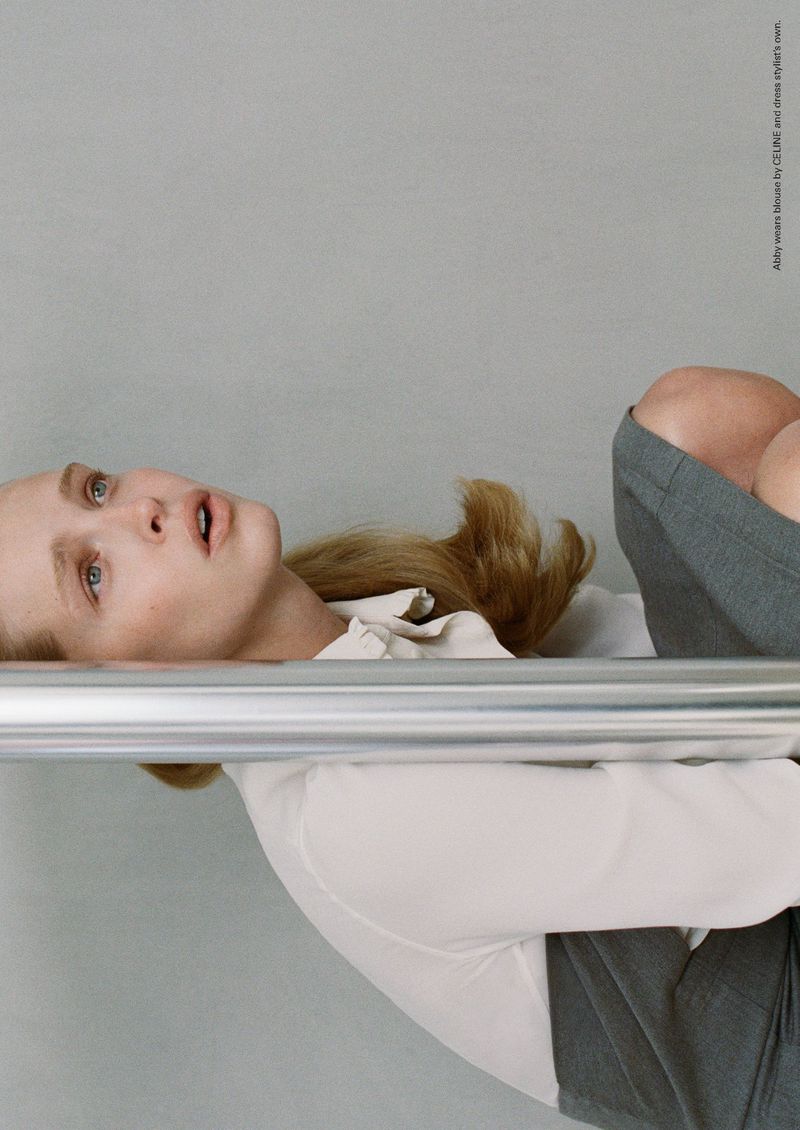 Model Abby Champion wearing a white collared shirt and grey skirt behind a horizontal chrome metal bar against a light grey studio background.