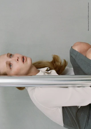 Model Abby Champion wearing a white collared shirt and grey skirt behind a horizontal chrome metal bar against a light grey studio background.