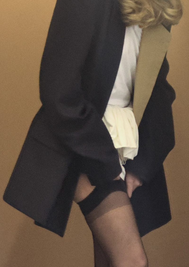 Woman poses indoors wearing a black blazer, white top, and black stockings against a neutral backdrop for fashion or beauty.