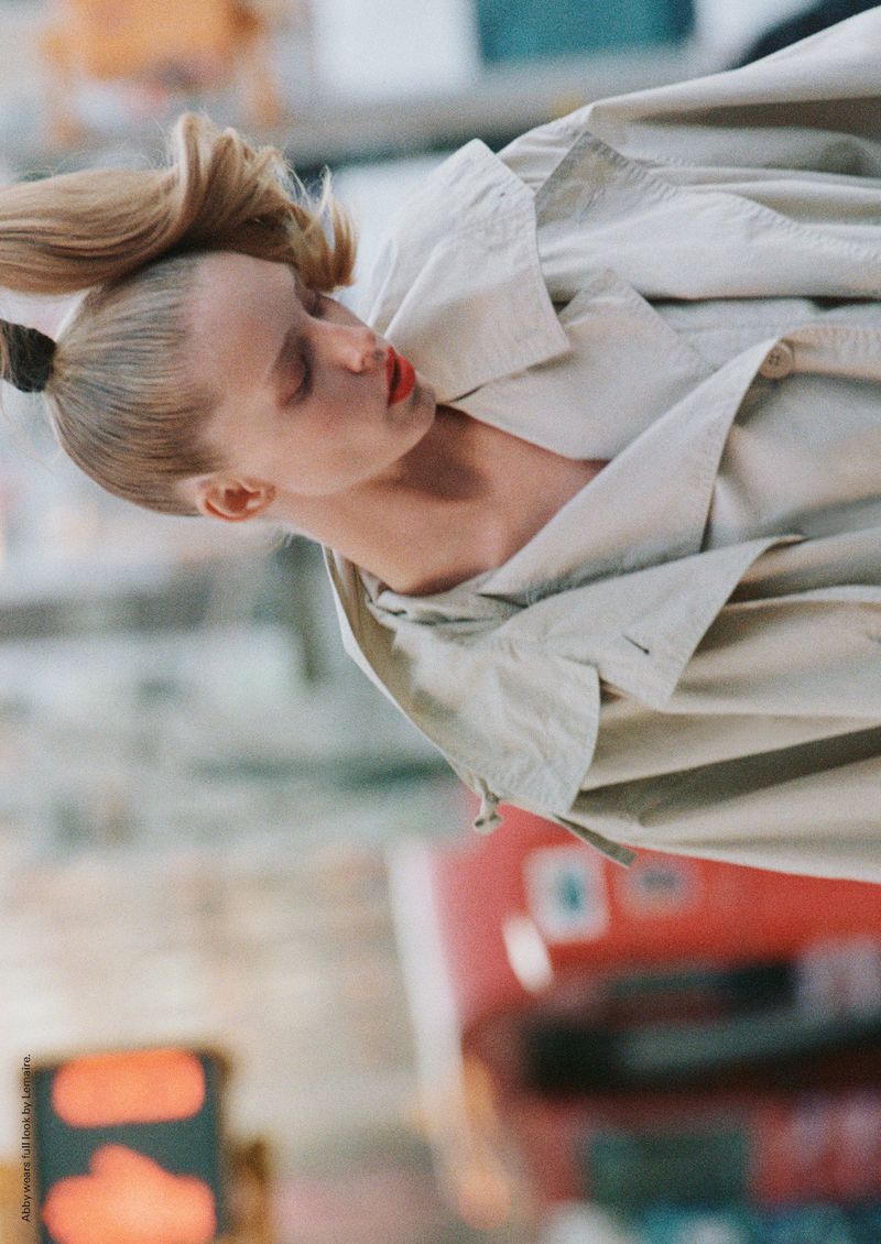 Blonde woman in trench coat with windblown hair, red lips, and neutral background, shot in a portrait orientation.
