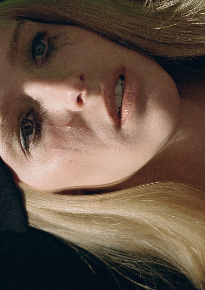 Close-up portrait of a blonde woman with light skin lying down, featuring striking makeup and dramatic lighting against a dark backdrop.