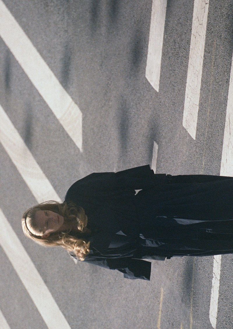 Woman with long blond hair walks across a gray asphalt crosswalk. She wears a black coat and the composition emphasizes verticality.