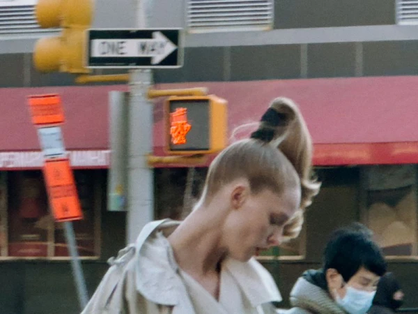 Woman in ponytail stands near traffic signal, buildings in New York City. She wears a tan coat on a city street in daylight.
