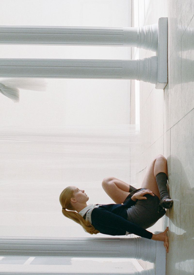 Indoor view of a young woman in a school uniform leaning back on her arms with her legs up, set against the backdrop of two white pillars.