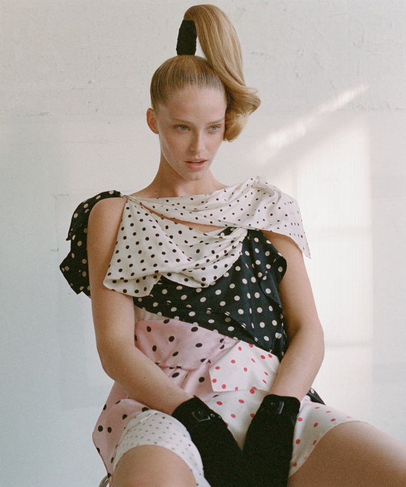 Fashionable adult woman with a blonde high ponytail poses while seated in a multi-patterned polka dot dress in front of a white wall.