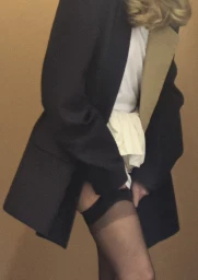 Model Abby Champion wearing an oversized black blazer and sheer stockings against a warm neutral studio backdrop.