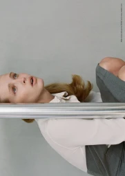 Model Abby Champion wearing a white collared shirt and grey skirt behind a horizontal chrome metal bar against a light grey studio background.