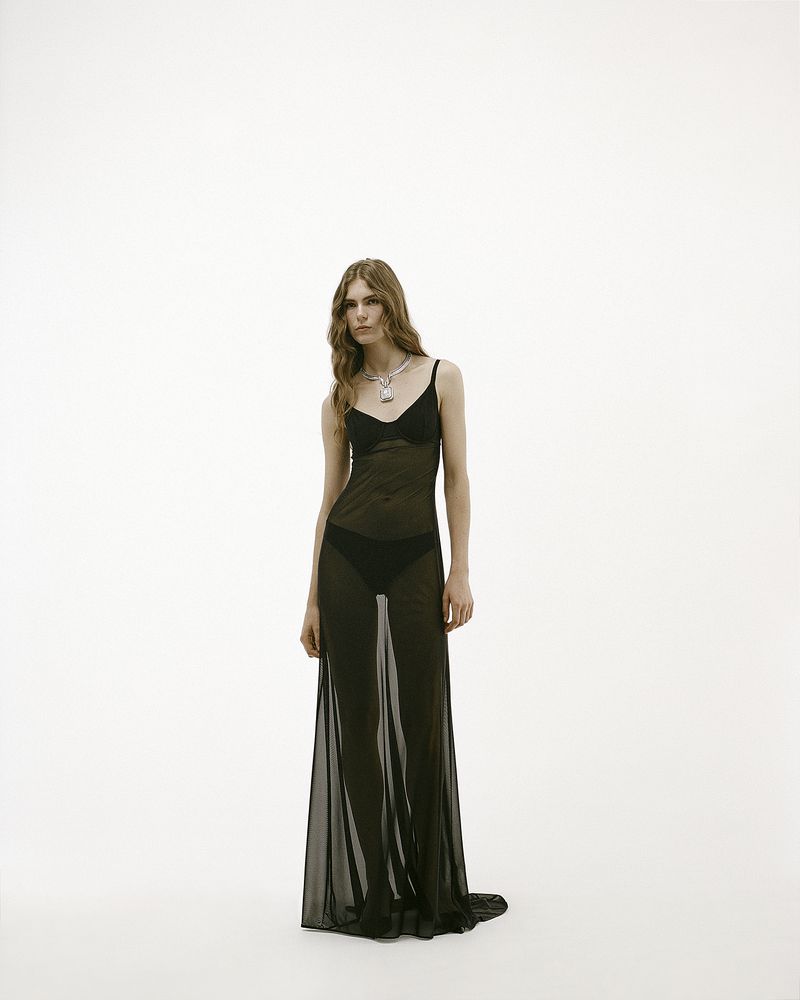 Model Anna Robinson poses in a sheer black floor-length dress and statement necklace against a white seamless background.