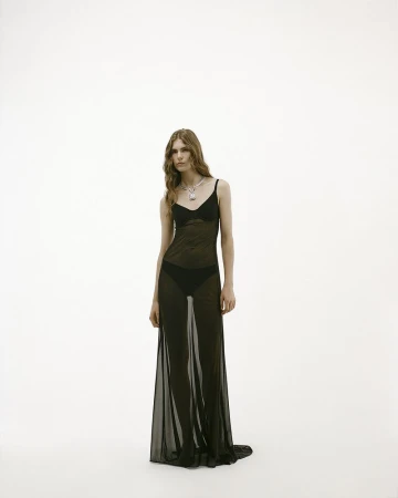 Model Anna Robinson poses in a sheer black floor-length dress and statement necklace against a white seamless background.