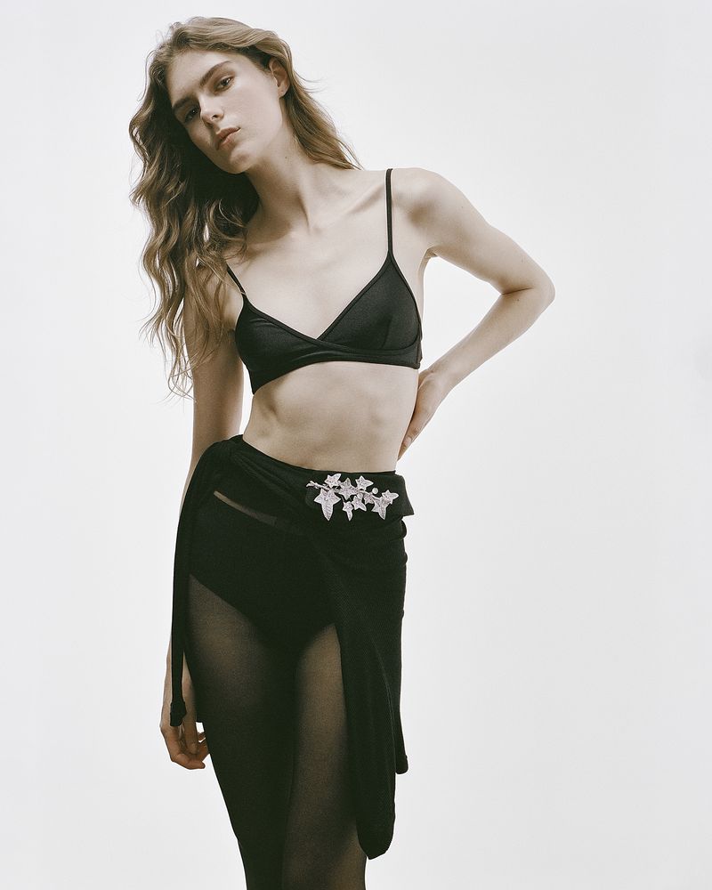 Model Anna Robinson posing in a black triangle bralette, sheer tights, and a wrap skirt with crystal brooch against a white backdrop.