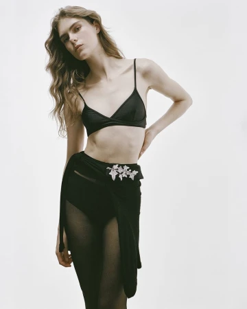 Model Anna Robinson posing in a black triangle bralette, sheer tights, and a wrap skirt with crystal brooch against a white backdrop.