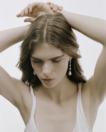 Model Anna Robinson with hands behind head wearing dangling diamond earrings and a white top against a white backdrop.
