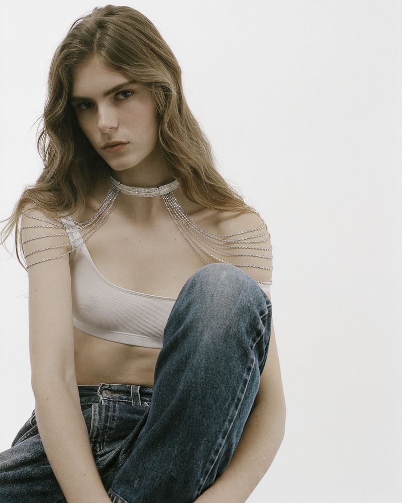 Anna Robinson seated in a pale crop top, draped rhinestone shoulder jewelry, and denim jeans against a white seamless background.