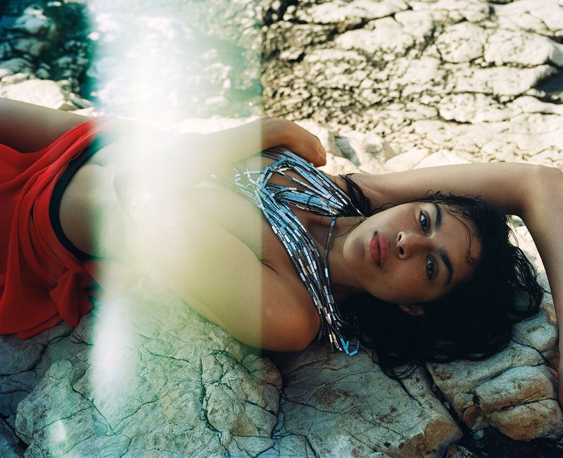 A woman with dark hair and a jeweled top is reclining on light-colored rocks near the ocean. She is wearing a red skirt, and the light is diffused.
